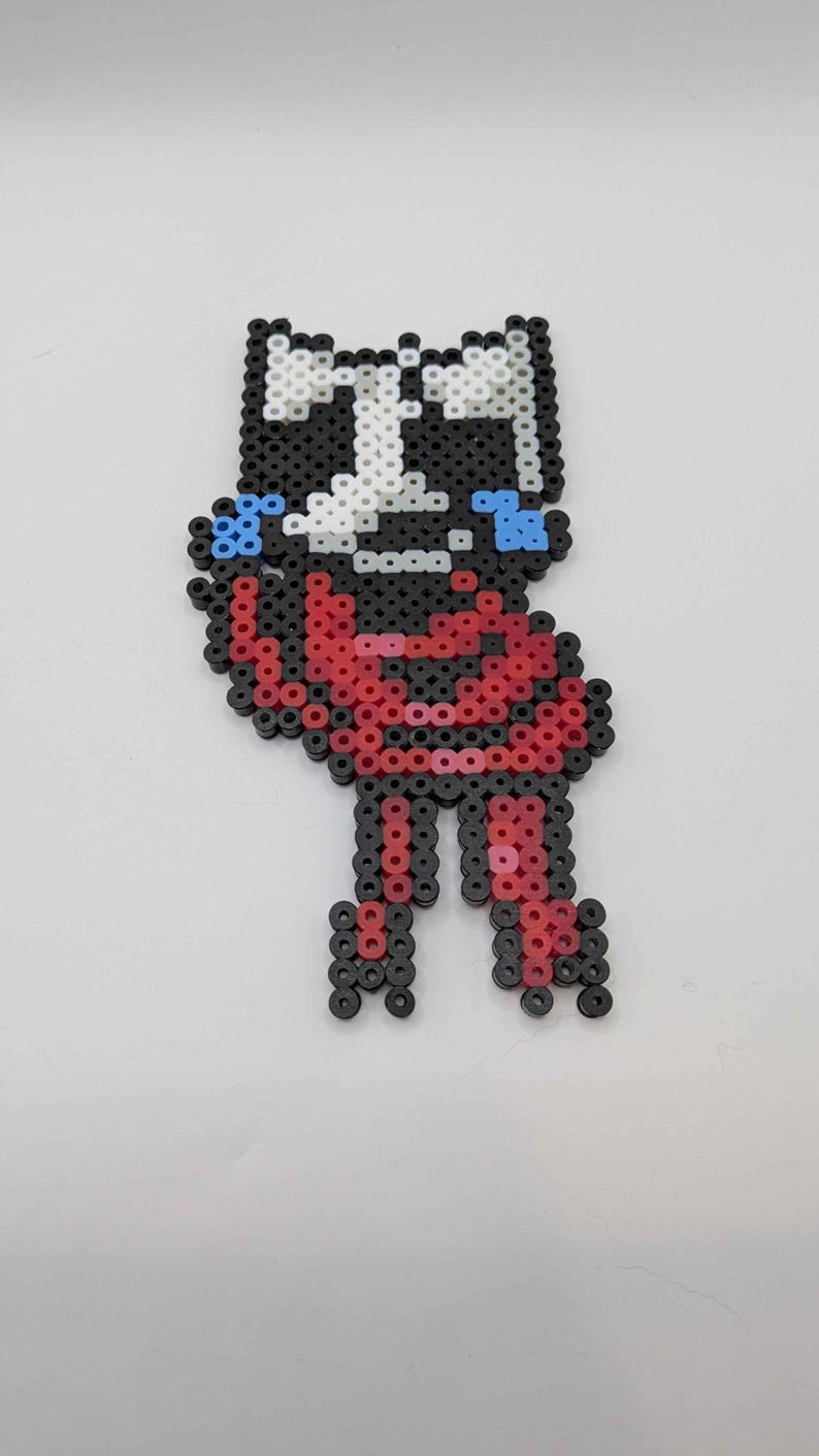 May include: A pixel art figure made from colorful plastic beads. The figure has a black and white head, red body, and black legs. Blue accents are on the figure's face. The figure is standing on a white surface.