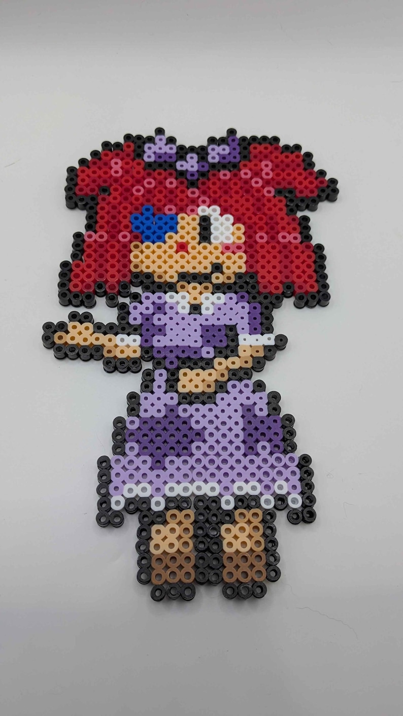 May include: A pixel art figure of a girl made from colorful plastic beads. The figure has red hair, a purple dress, and brown shoes. The figure is set against a white background.