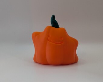 Puede incluir: Una figura de color naranja brillante con forma de calabaza, acabado mate liso y un tallo verde oscuro en la parte superior. La figura presenta una cara sonriente sutilmente grabada con dos ojos pequeños y una boca curva en el centro.