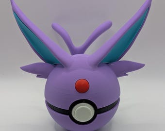 May include: A spherical purple toy resembling a creature with two large, pointed ears featuring teal inner accents. The object has a central black band, a white circular button, and a small red circular detail positioned above the center.