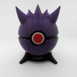 Puede incluir: Escultura morada impresa en 3D inspirada en una Poké Ball. El objeto esférico presenta un botón rojo y detalles negros, con protuberancias puntiagudas en la parte superior. Descansa sobre un soporte negro de tres patas, sobre un fondo blanco.