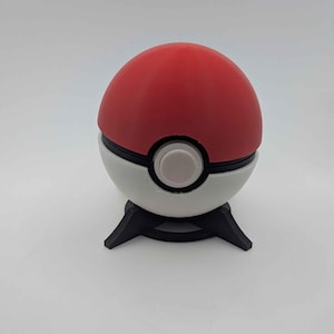 Può includere: Una replica di Poké Ball rossa e bianca su una base nera. La palla ha un centro bianco con un bottone grigio. La base ha tre gambe. La palla è sferica.