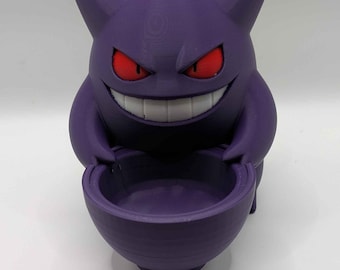 Puede incluir: Una figura del personaje Gengar de color morado con orejas puntiagudas, púas en la cabeza, ojos rojos y una amplia sonrisa blanca. La figura sostiene un pequeño recipiente morado en forma de cuenco entre sus manos.