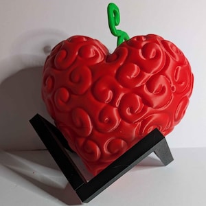 May include: A red, heart-shaped object with a textured surface and a green stem, displayed on a black stand. The object has a unique, swirling pattern across its surface. The stand is a simple, angular design.