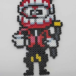 May include: A pixel art figure made from colorful plastic beads. The figure has a red and white face, a black body, and yellow accents. The figure is standing with its arms at its sides.
