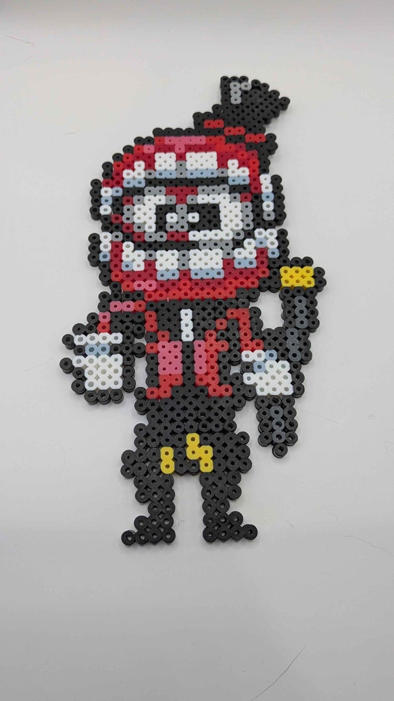 May include: A pixel art figure made from colorful plastic beads. The figure has a red and white face, a black body, and yellow accents. The figure is standing with its arms at its sides.
