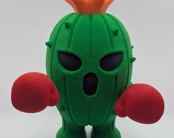 Puede incluir: Una figura verde con forma de cactus, con ojos negros y expresión de sorpresa. Lleva guantes de boxeo rojos y una corona naranja. Un artículo decorativo caprichoso.
