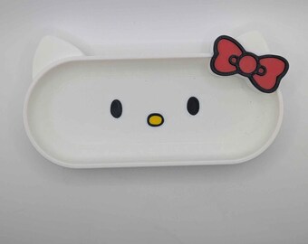 Puede incluir: Una bandeja blanca de forma ovalada con un diseño de cara de gato de dibujos animados. La bandeja presenta ojos negros, una nariz amarilla y un lazo rojo. La bandeja tiene dos orejas blancas a los lados.