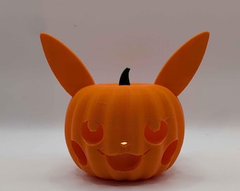 Puede incluir: Un recipiente decorativo naranja con forma de calabaza que presenta dos orejas altas y puntiagudas y una cara tallada con mejillas circulares, ojos almendrados y una boca ancha y sonriente. Un pequeño tallo negro se asienta encima y una luz brilla desde el interior.
