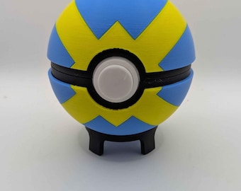 Puede incluir: Una esfera con un patrón geométrico azul y amarillo, que presenta una banda negra central y un botón circular blanco en la parte frontal. El objeto se apoya sobre un pequeño soporte negro y muestra un acabado mate y liso en su superficie.