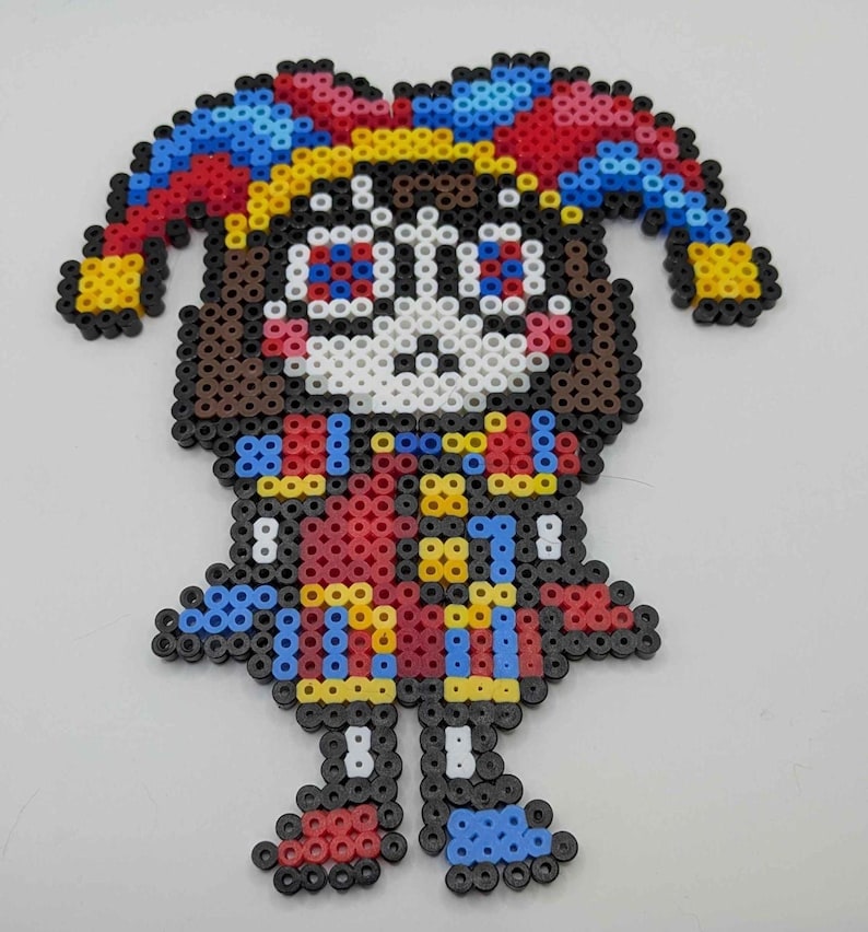 May include: A colorful jester figure crafted from fused plastic beads. The jester has a white face, brown hair, and wears a red and blue outfit with yellow accents. The jester's hat is a vibrant mix of red, blue, and yellow.