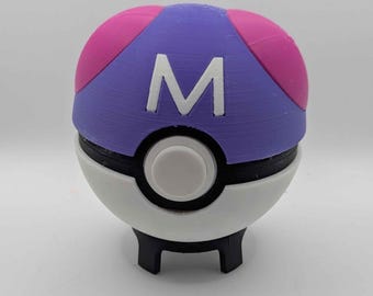 Puede incluir: Una réplica esférica de Master Ball con una mitad superior morada con detalles en rosa y una mitad inferior blanca. Una letra M blanca está centrada en la parte superior y un botón circular blanco se sitúa en el centro sobre un soporte negro.