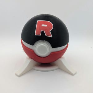 Könnte beinhalten: Eine schwarz-rot-weiße Pokéball-Replik mit rotem "R"-Logo, auf einem weißen Ständer. Der Ball ist in drei Abschnitte unterteilt: eine schwarze Oberseite, eine weiße Mitte und ein roter Boden. Der Ständer hat drei weiße Beine.