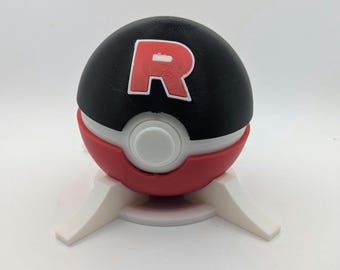 May include: A black, red, and white Poké Ball replica with a red "R" logo, displayed on a white stand. The ball is split into three sections: a black top, a white center, and a red bottom. The stand has three white legs.