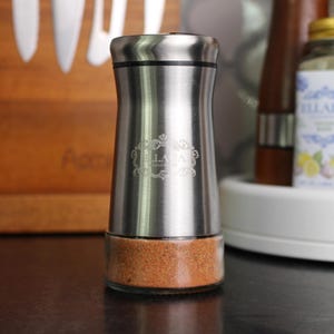 May include: A stainless steel spice grinder with a clear glass base filled with a reddish-brown spice blend. The grinder has a silver top and a decorative logo. The grinder is designed for kitchen use.