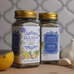 May include: Two glass jars of Ellada Tzatziki Blend seasoning with gold lids. The labels feature blue floral designs, lemons, and garlic. The net weight is 3.48 oz (98.79g). A lemon half and garlic cloves are in the foreground.