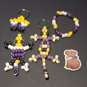 May include: A collection of handmade beaded accessories in non-binary pride colors. Includes a star-shaped keychain, a lizard-shaped keychain, a bracelet, a lizard-shaped figure, and a capybara sticker. The beads are yellow, white, purple, and black.