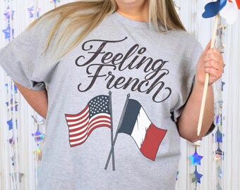 Puede incluir: Una camiseta gris jaspeado con la frase "Feeling French" en letra cursiva. La camiseta presenta banderas cruzadas de Estados Unidos y Francia. Un molinillo rojo, blanco y azul se sostiene en la mano.