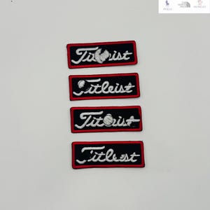 Könnte beinhalten: Vier gestickte Aufnäher mit dem Wort "Titleist" in weißer Schrift auf einem schwarzen rechteckigen Hintergrund mit rotem Rand. Zwei Aufnäher zeigen ein Golfball-Design.