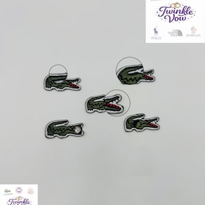 May include: A collection of embroidered crocodile logo patches in green, with red detailing. The patches vary in size and are designed for application on clothing or accessories. The image shows several patches.