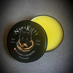 May include: A black tin of NawaKitty rope butter or balm with a black cat illustration. The tin is open, revealing a yellow balm. Text includes "Rope Butter", "Rope Balm", and "Do Not Ingest".