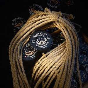 May include: A bundle of tan rope with several small, round, black containers. The containers have a white and black cat logo and the text "NawaKitty". The text "Rope Butter" and "Rope Balm" are also visible. The background is dark.