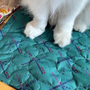 May include: A quilted fabric mat in a teal green color featuring a repeating pattern of small dark blue and red geometric squares. The mat has a stitched circular design throughout the surface and is partially covered by white animal fur.