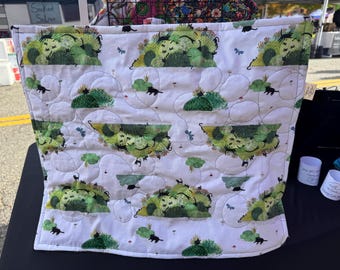 May include: A square quilted fabric bag featuring a white background with a repeating pattern of green grassy mounds, small black cats, and tiny blue butterflies. The fabric has visible machine stitching in a swirling, rounded pattern.