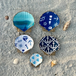 May include: Five decorative seashells with nautical designs on a sandy surface. The shells feature painted blue gradients and patterns with anchors, sailboats, and nautical symbols. The shells are painted with gold edges.