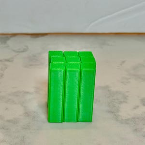 Pode incluir: Um cubo verde brilhante, impresso em 3D, composto por nove prismas retangulares. O cubo está ligeiramente descentrado sobre um fundo neutro. A cor verde vibrante é consistente em todo o objeto.