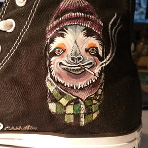 May include: Black canvas high-top sneaker with a hand-painted sloth design. The sloth wears a purple and white striped beanie, a green and black plaid shirt, and smokes a cigarette. White laces and a white rubber sole complete the shoe.