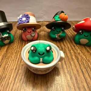 May include: A collection of whimsical frog figurines. One frog wears a top hat, another a witch's hat with a pumpkin, and another a mushroom cap. A frog sits in a teacup. The frogs are green, pink, and white.