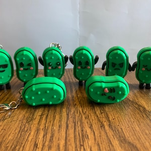 May include: A collection of green pickle-shaped keychains with various facial expressions. Some have arms and legs. The keychains are made of a rubbery material and have a textured surface. The keychains are on a wooden surface.