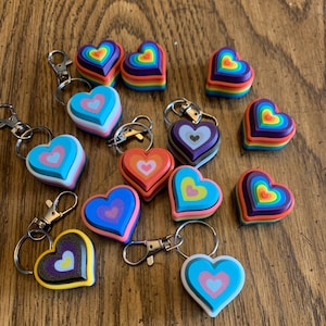 May include: A collection of heart-shaped keychains in various colors, including blue, pink, yellow, and rainbow. Each keychain has a metal clasp and ring. The hearts are layered with different colors, creating a 3D effect. The keychains are arranged on a wooden surface.