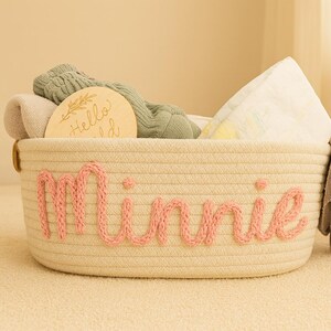 May include: A cream-colored woven rope basket featuring the name Minnie embroidered in pink yarn on the front. The basket is filled with folded textiles, including a sage green garment and a round wooden disc engraved with the text Hello World.