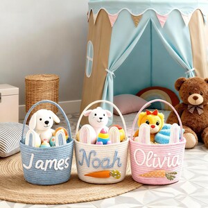Puede incluir: Tres cestas redondas de cuerda trenzada con asas, en colores azul claro, crema y rosa claro. Cada cesta presenta un nombre personalizado en letra cursiva blanca o azul, orejas de conejo de fieltro y zanahorias decorativas de fieltro.