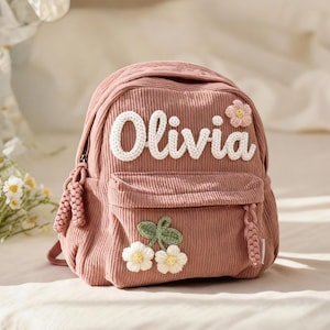 Puede incluir: Una mochila de pana rosa polvoriento con el nombre "Olivia" en letras blancas de ganchillo. La mochila tiene un bolsillo delantero y está decorada con flores y cerezas de ganchillo. La bolsa tiene un asa superior y dos borlas laterales.