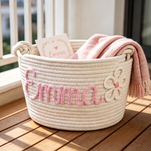 May include: A round, off-white rope basket with a pink knitted name "Emma" and a flower applique. The basket contains a pink blanket and a card with the name "Emma".
