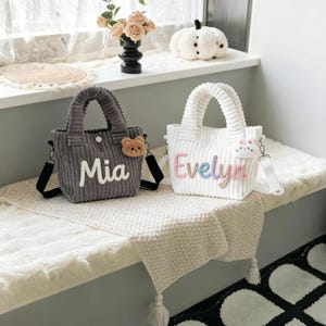 Puede incluir: Dos bolsos tote de pana, uno gris y otro blanco, se muestran uno al lado del otro. El bolso gris tiene el nombre "Mia" bordado en blanco y un charm de oso. El bolso blanco tiene el nombre "Evelyn" bordado en colores pastel y un charm de gato. Ambos tienen asas y correas para el hombro.