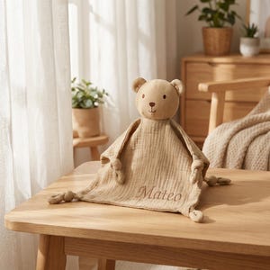 Puede incluir: Una manta de seguridad para bebé de color beige con una cabeza de oso de peluche y esquinas anudadas. El nombre "Mateo" está bordado en la manta. El juguete de peluche está diseñado para la comodidad y la seguridad.