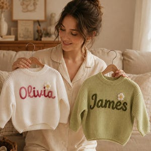 Op de afbeelding: Twee kindertruien, één crème en één saliegroen, worden getoond op houten kleerhangers. De crèmekleurige trui heeft de naam "Olivia" geborduurd in roze, en de groene trui heeft "James" in donkergroen. Beide hebben een madeliefje.