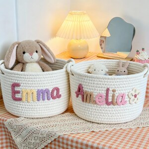 Puede incluir: Dos cestas de almacenamiento de cuerda blanca con asas, cada una bordada con un nombre en letras coloridas. Una dice "Emma" y la otra "Amelia". Cada cesta contiene un peluche. Las cestas están sobre una superficie a cuadros.