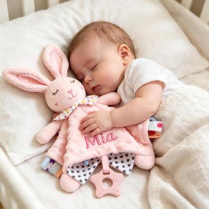 Puede incluir: Un bebé dormido abraza una manta de seguridad rosa con forma de conejito. La manta tiene una superficie texturizada y un estampado de lunares. El nombre "Mia" está bordado en la manta. Un mordedor rosa en forma de estrella está adjunto.