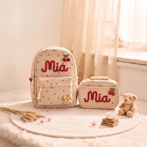 Puede incluir: Una mochila de pana beige y una bolsa de almuerzo rectangular a juego, ambas con un estampado de pequeñas cerezas rojas y el nombre Mia bordado en rojo. La mochila incluye un pequeño parche de cara de tigre y un dije de cereza en la parte superior.