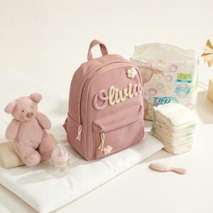 May include: A pink corduroy backpack with the name "Olivia" in cream script. The backpack has a small front pocket and flower accents. A pink teddy bear, baby bottle, nappies, and a hairbrush are also in the image.