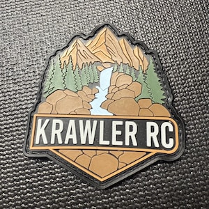 以下が含まれることがあります： 滝、木、岩のある山の風景を描いたパッチ。パッチは黒い縁取りで、茶色とタンの色使いです。「KRAWLER RC」の文字が白で表示されています。