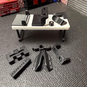 Puede incluir: Una colección de piezas negras impresas en 3D para coches RC, incluyendo soportes y componentes del chasis, dispuestas sobre una mesa blanca. Las piezas son de un material negro mate. El texto "Krawler RC" es visible en una de las piezas.