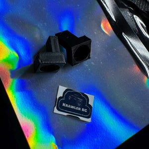 May include: Two black, 3D-printed RC car parts with circular openings sit on a reflective, iridescent surface. A small sticker with the text "KRAWLER RC" and a graphic of a vehicle is also present. The parts have a textured appearance.