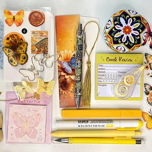 May include: A collection of stationery items including a butterfly-themed notebook, a patterned pen with a gold tassel, yellow highlighters, butterfly-shaped paper clips, a book review notepad, a round floral tin, and various butterfly stickers.