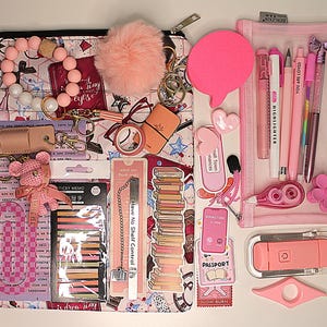 May include: A selection of pink and peach stationery and accessories, including pens, stickers, a pom-pom, a bookmark, and a reading light. The items are arranged on a white surface, with a patterned pouch and a clear pencil case. The image includes the text "YOU ARE LOVED".
